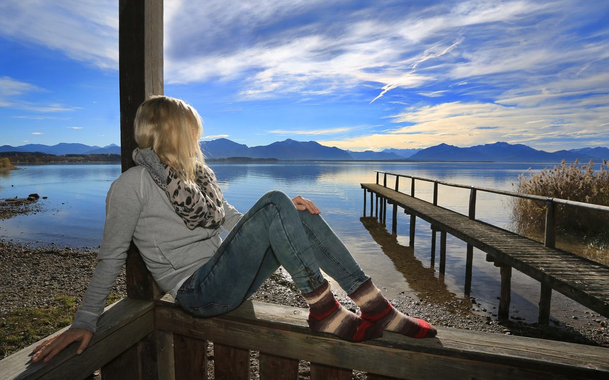 Andrea Wittmann sitzt am Bootshaus-Geländer, Blick über den Chiemsee zur Alpenkette
