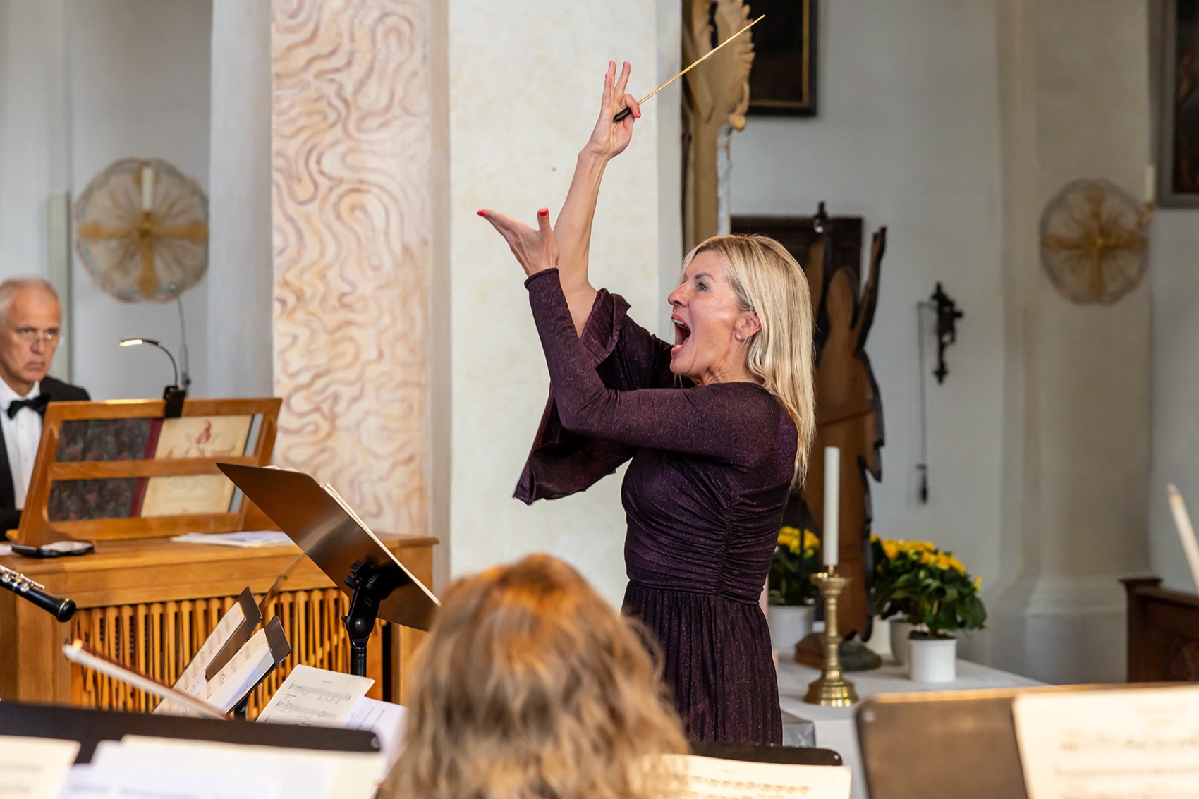 Andrea Wittmann conducting in a church, baton raised, both arms in motion