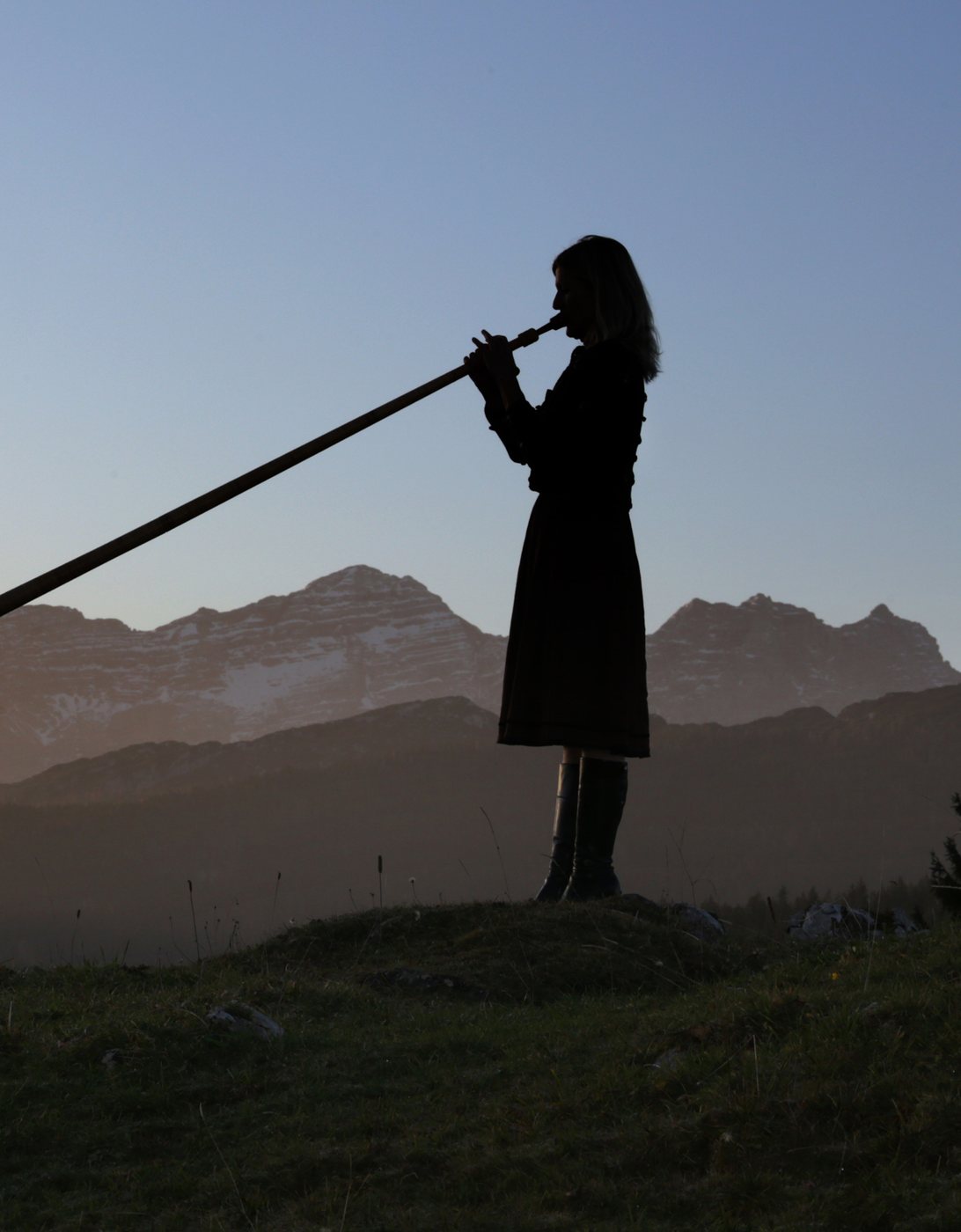 Andrea Wittmann spielt Alphorn in der Dämmerung, als Silhouette vor der Bergkette